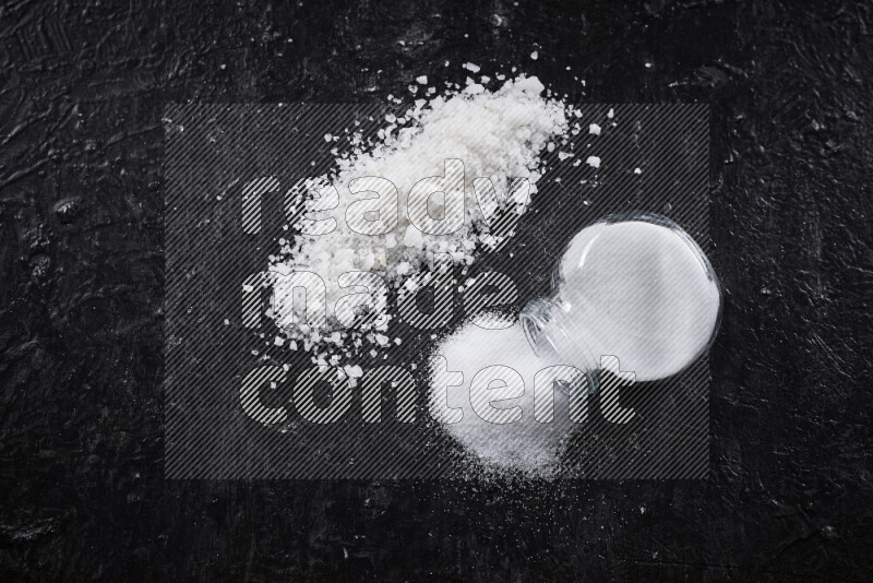 A glass jar full of table salt with some sea salt crystals beside it on a black background