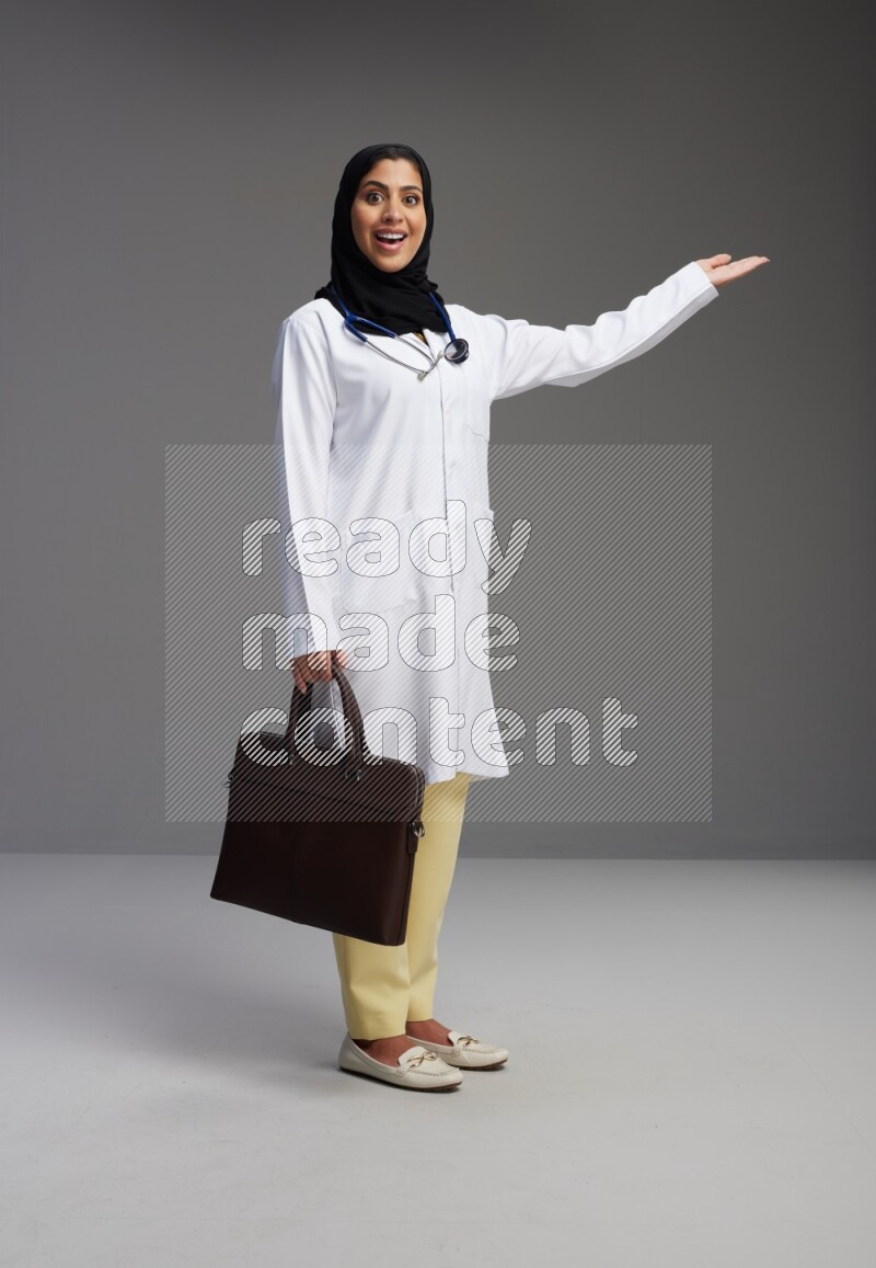Saudi woman wearing lab coat with stethoscope standing holding bag on Gray background