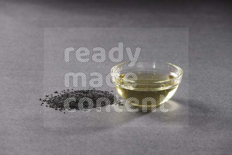 A glass bowl full of black seeds oil and black seeds beside it on a black flooring