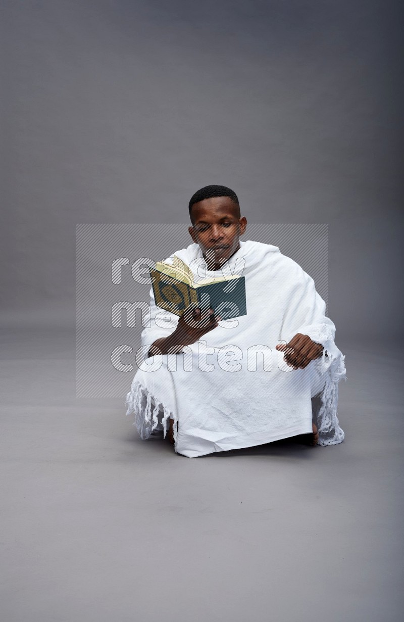 A man wearing Ehram sitting on floor reading quran on gray background