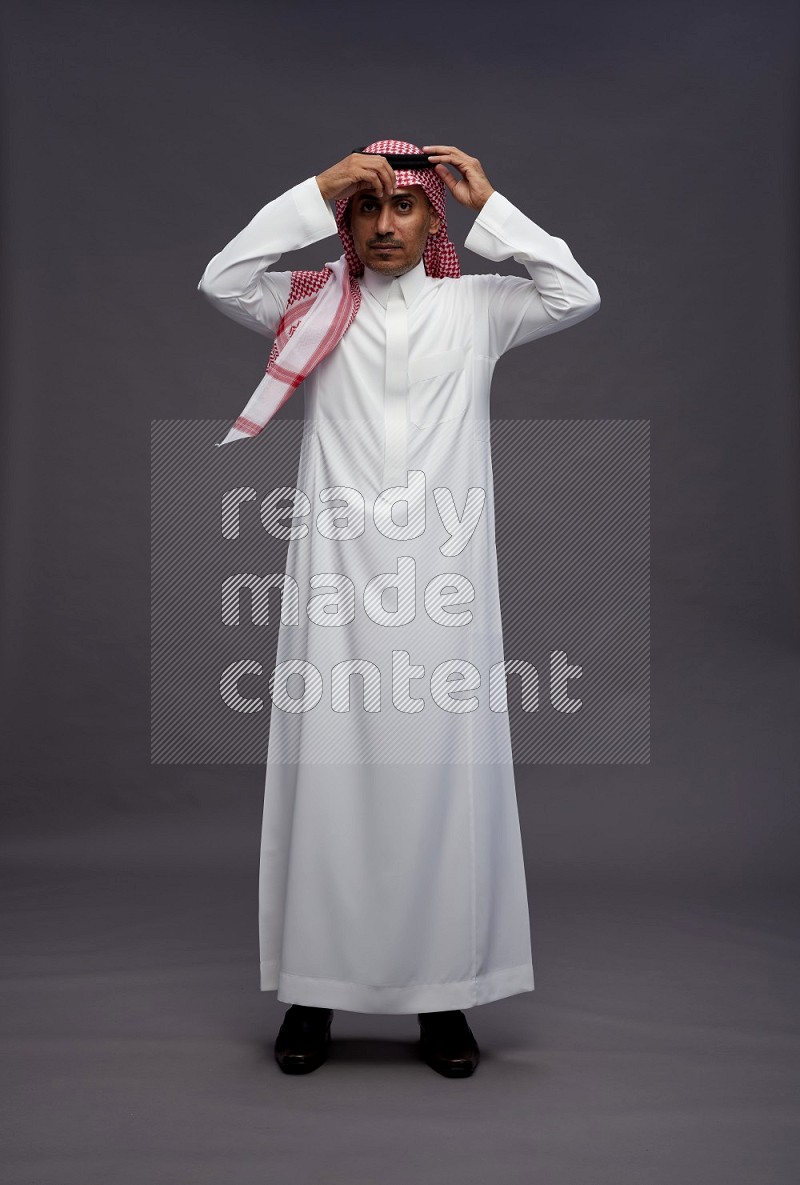 Saudi man wearing thob and shomag standing hands behind head on gray background