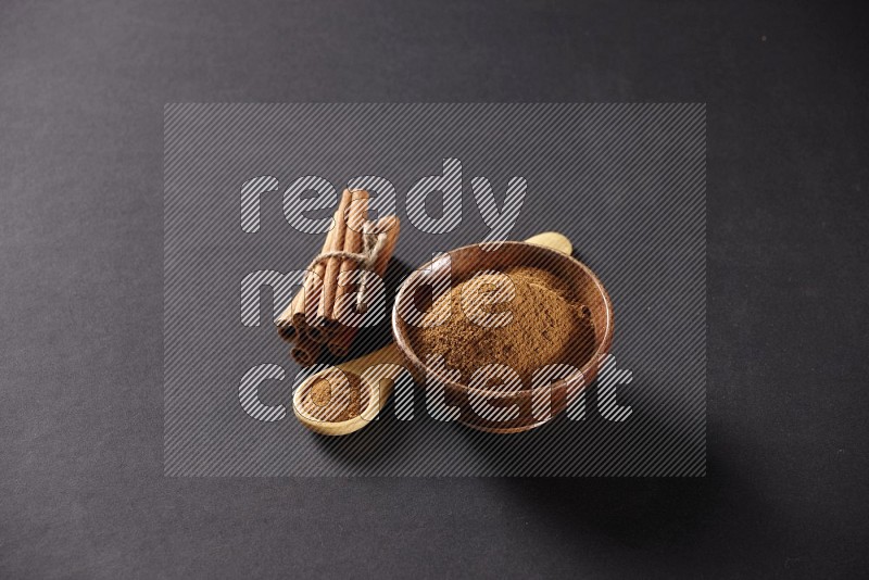 Cinnamon sticks stacked and bounded beside a wooden bowl full of cinnamon powder and a wooden spoon full of powder on black background