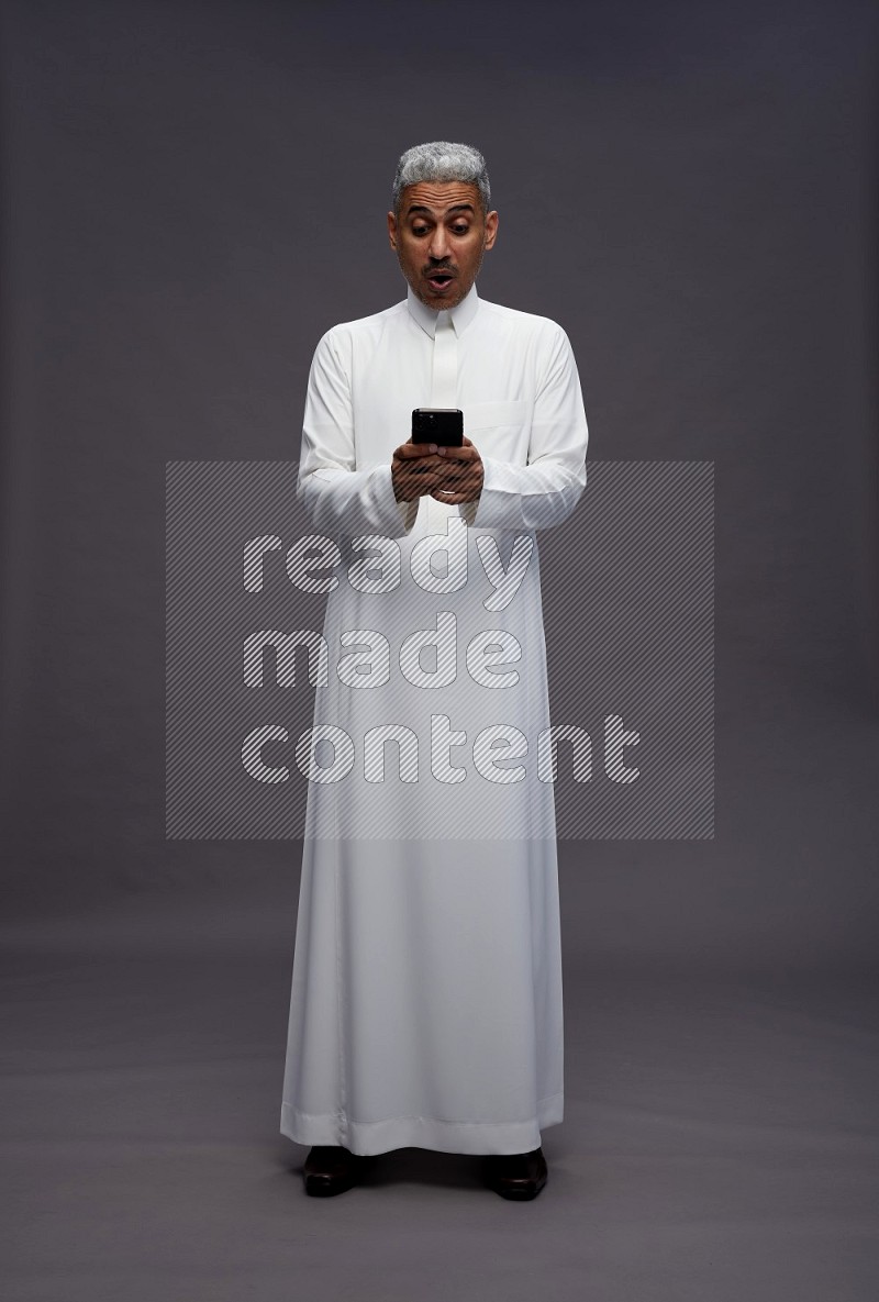 Saudi man wearing thob standing texting on phone on gray background