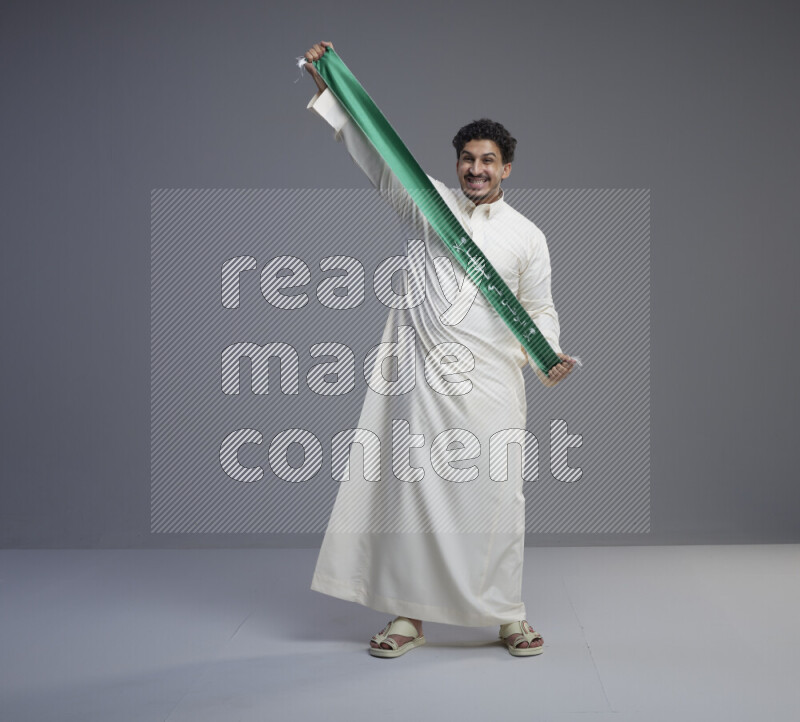 A Saudi man standing wearing thob holding Saudi flag scarf on gray background