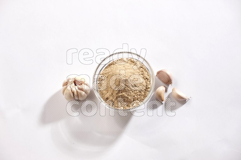 A glass bowl full of garlic powder with garlic bulb and some cloves beside it on a white flooring