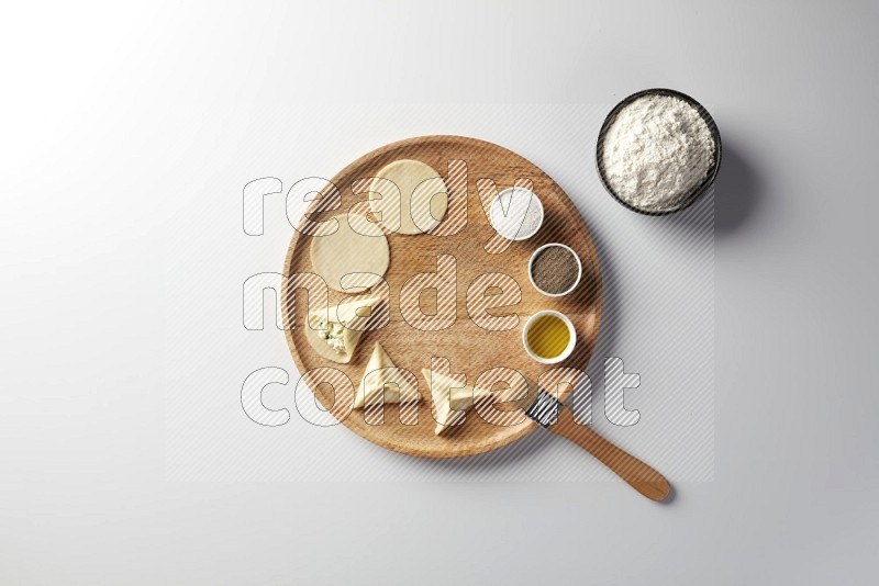 two closed sambosas and one open sambosa filled with cheese while flour, salt, black pepper and oil with oil brush aside in a wooden dish on a white background