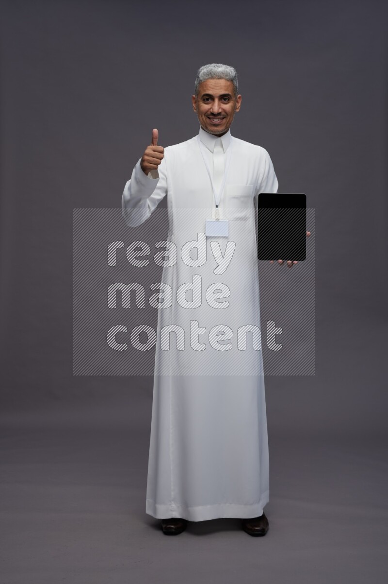 Saudi man wearing thob with neck strap employee badge standing showing tablet to camera on gray background