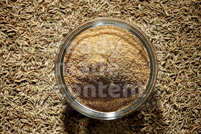 A glass bowl full of cumin powder surrounded by cumin seeds on black flooring