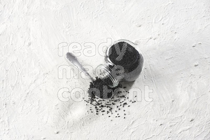 A glass spice jar and a metal spoon full of black seeds and jar is flipped with fallen seeds on a textured white flooring in different angles