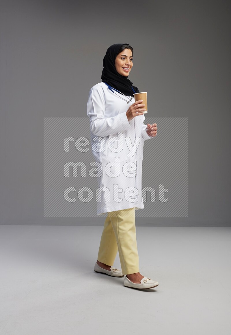 Saudi woman wearing lab coat with stethoscope standing holding paper cup on Gray background