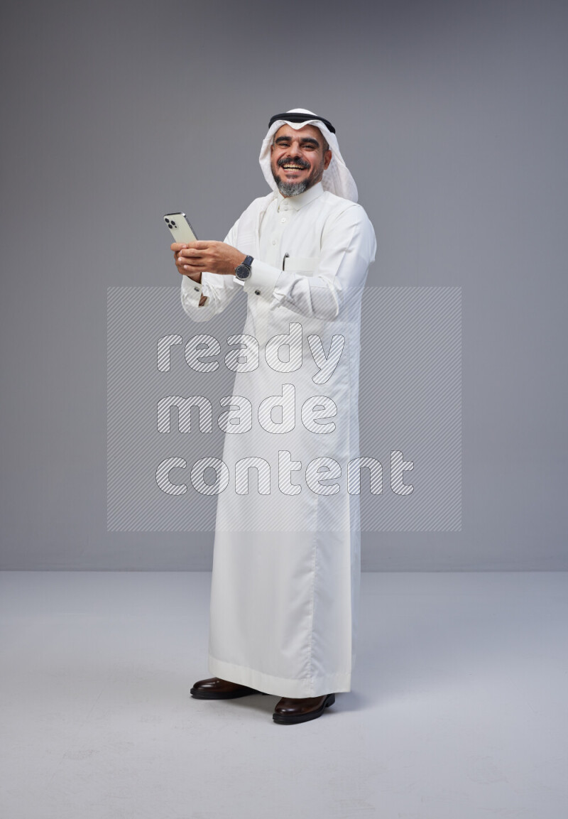 Saudi man Wearing Thob and white Shomag standing texting on phone on Gray background