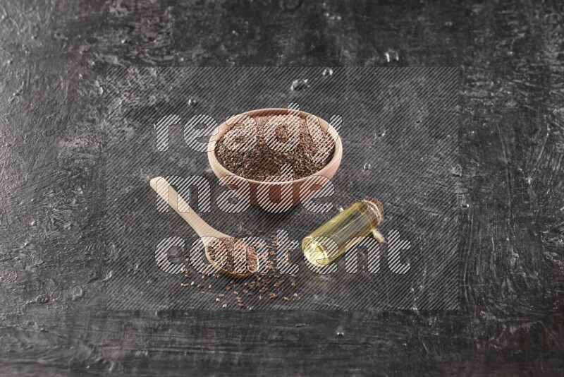 A wooden bowl and spoon full of flaxseeds and a glass bottle of flaxseeds oil on a textured black flooring