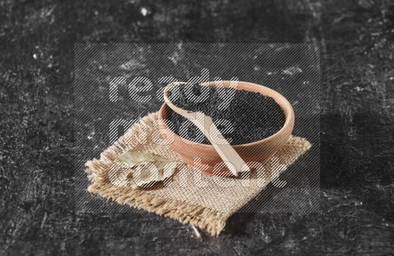 A wooden bowl full of black seeds with wooden spoon full of the seeds on it on a burlap fabric on a textured black flooring