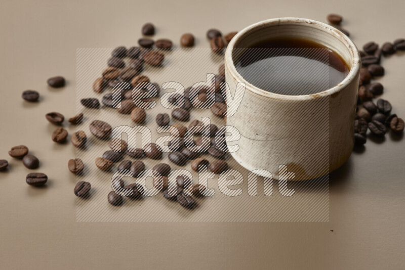 A beige pottery cup of coffee surrounded by roasted coffee beans on beige background