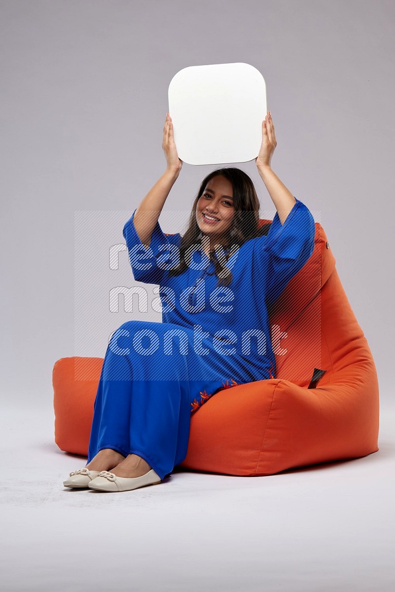A Woman sitting on an orange beanbag wearing Jalabeya holding a social media sign