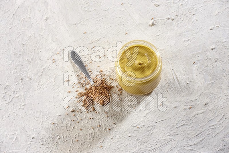 A glass jar full of mustard paste and mustard seeds in a metal spoon on a textured white flooring in different angles
