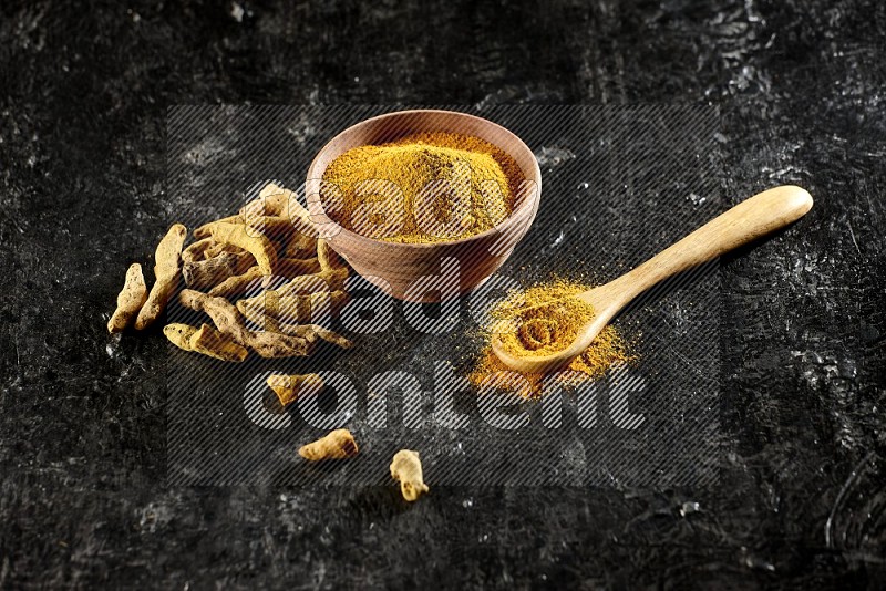 A wooden bowl and a wooden spoon full of turmeric powder with dried turmeric fingers on textured black flooring