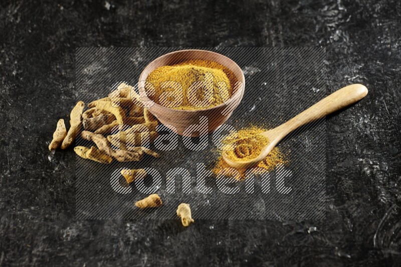 A wooden bowl and a wooden spoon full of turmeric powder with dried turmeric fingers on textured black flooring