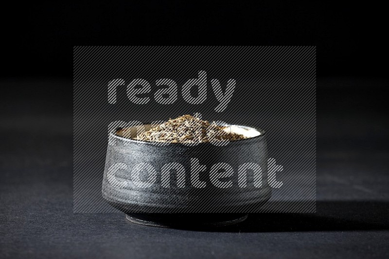 A black pottery bowl full of cumin seeds on a black flooring