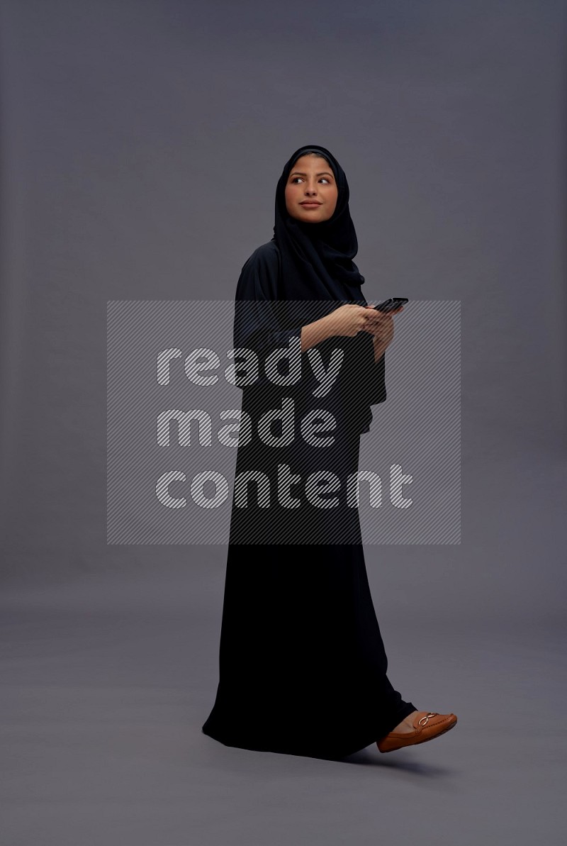 Saudi woman wearing Abaya standing texting on phone on gray background