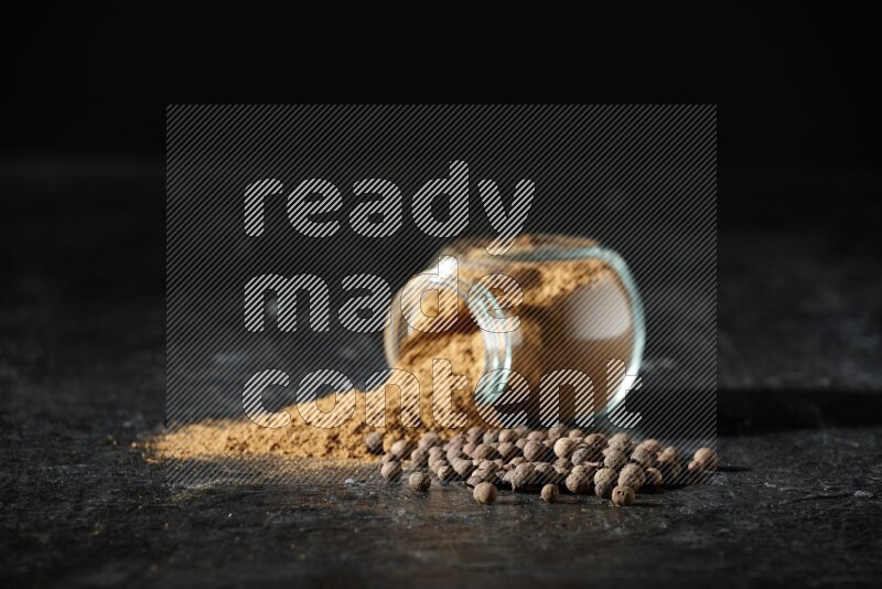 A flipped glass spice jar full of allspice powder and powder spilled out of it with whole balls on a textured black flooring