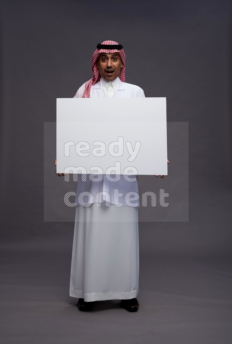 Saudi man wearing thob with lab coat and shomag with pocket employee badge standing holding board on gray background