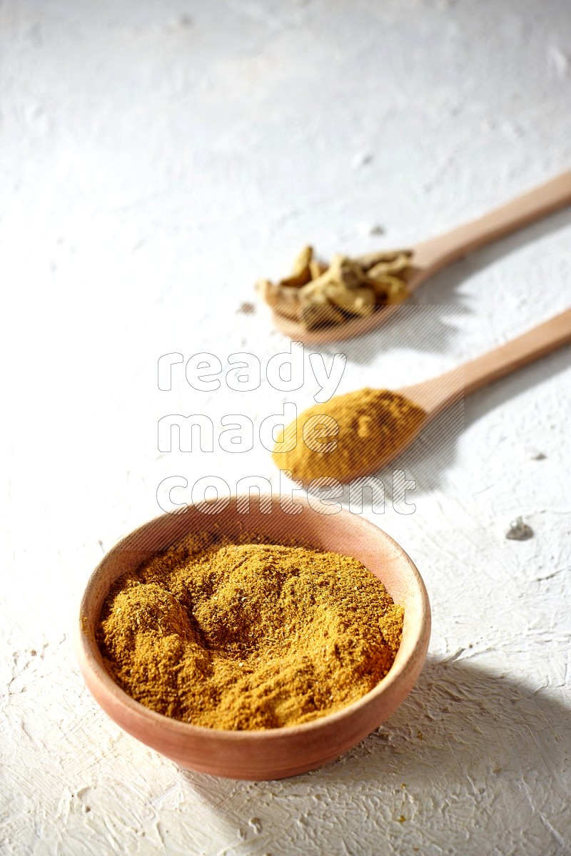 A wooden bowl full of turmeric powder and 2 wooden spoons full of dried turmeric whole finger and turmeric powder on textured white flooring