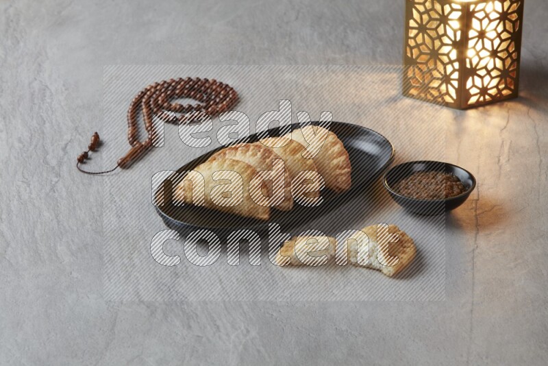 Four fried sambosas in an oval shaped black plate, beside a cut cheese sambosa, a brown misbaha and a golden lantern on a gray background