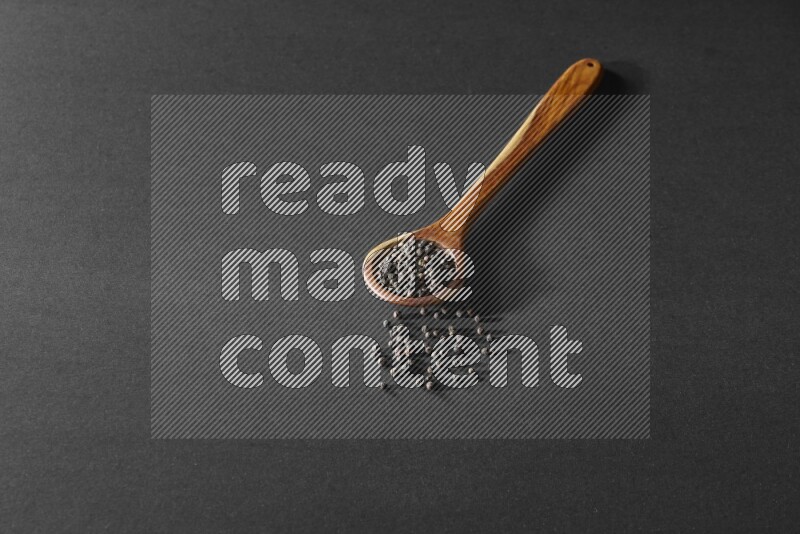 A wooden ladle full of black pepper beads on a black flooring