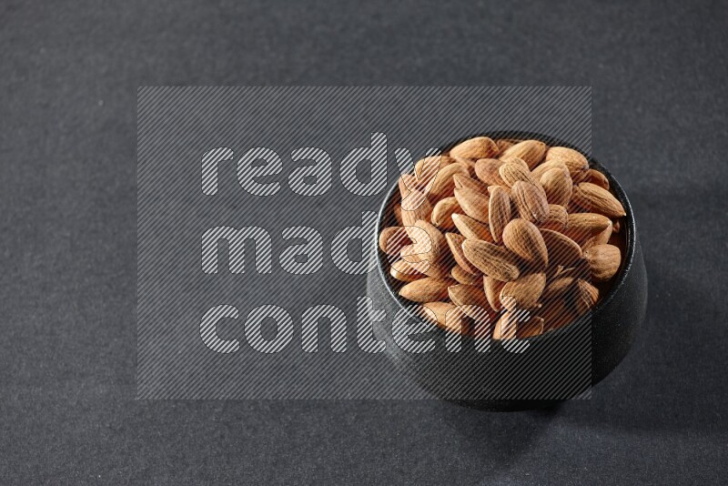 A black pottery bowl full of peeled almonds on a black background in different angles