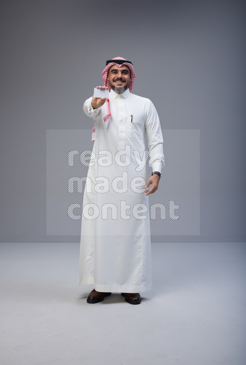 Saudi man Wearing Thob and red Shomag standing holding ATM card on Gray background