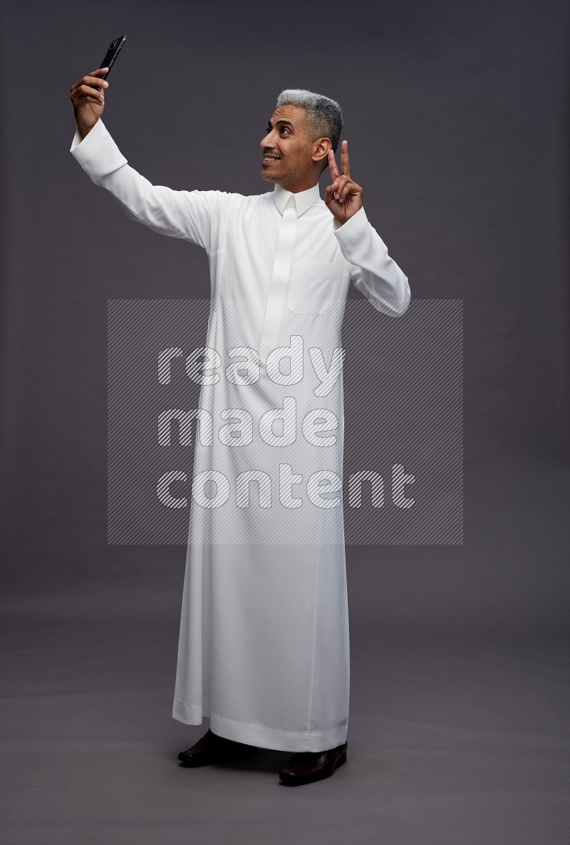 Saudi man wearing thob standing taking selfie on gray background