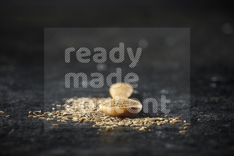 A wooden spoon full of cumin powder and cumin seeds spreaded on textured black flooring