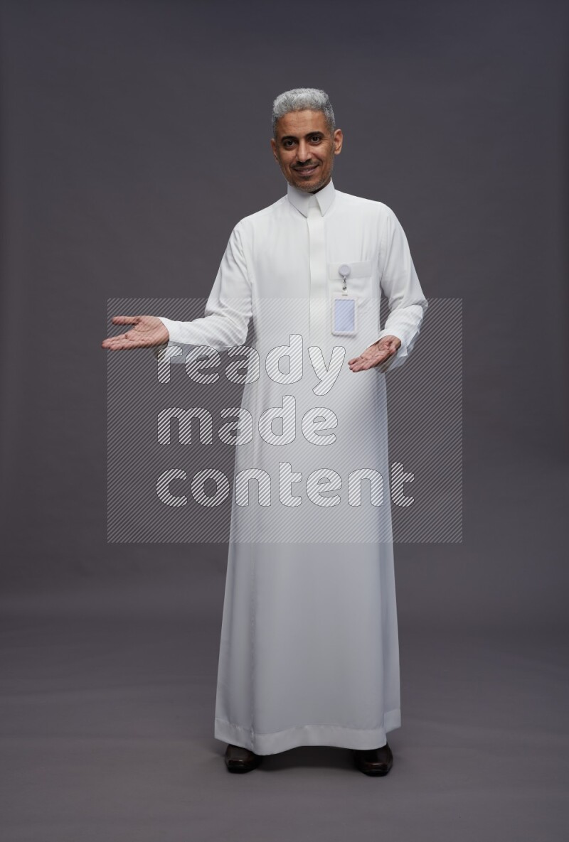 Saudi man wearing thob with pocket employee badge standing interacting with the camera on gray background