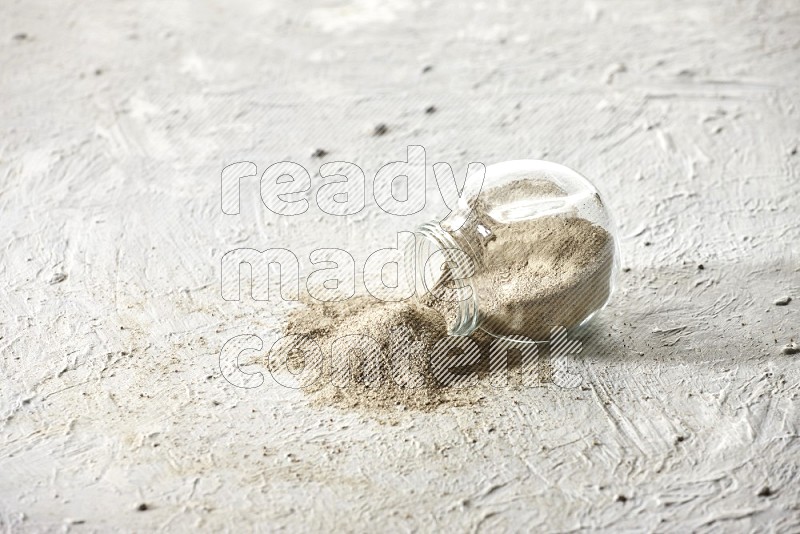 A flipped herbal glass jar full of white pepper powder with spilled powder on textured white flooring