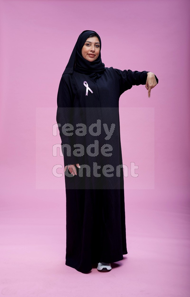 Saudi woman wearing pink ribbon on Abaya standing interacting with the camera on pink background