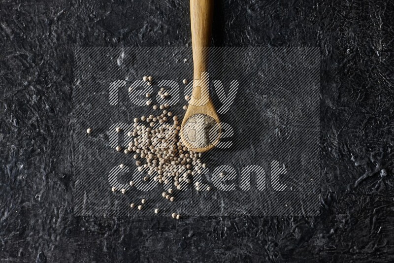 A wooden spoon full of white pepper powder with white pepper beads on textured black flooring