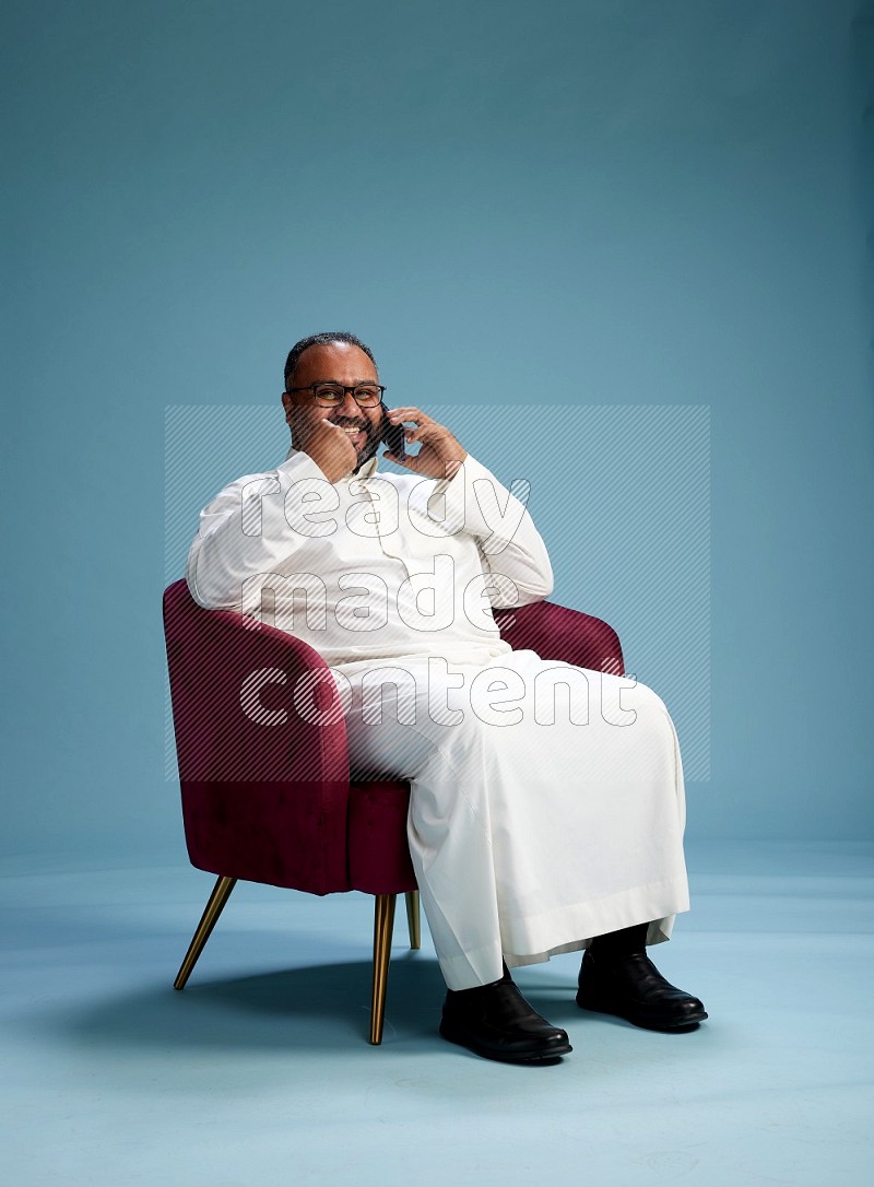 Saudi Man without shimag sitting on chair talking on phone on blue background