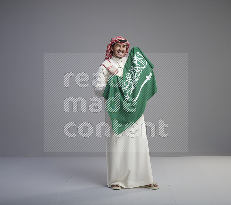 A saudi man standing wearing thob and red shomag holding big saudi flag on gray background