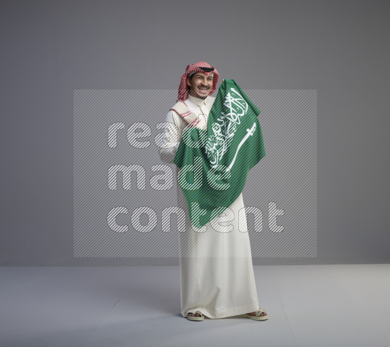 A saudi man standing wearing thob and red shomag holding big saudi flag on gray background