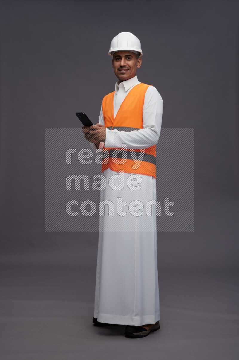 Saudi man wearing thob with engineer vest standing texting on phone on gray background
