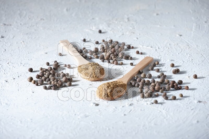 Wooden spoons full of allspice powder and allspice whole balls spreaded on a textured white flooring