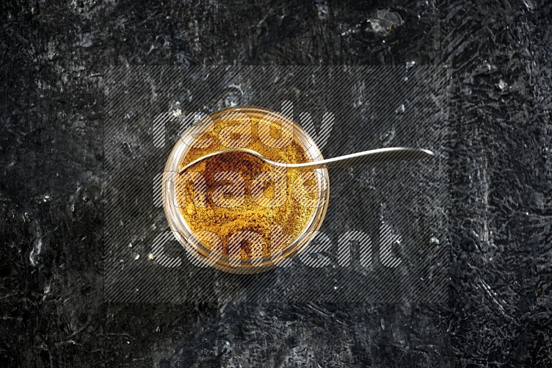 A glass jar and a metal spoon full of turmeric powder on a textured black flooring