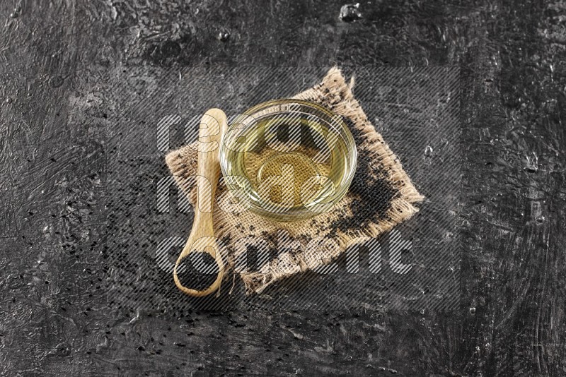 A glass bowl full of black seeds oil and wooden spoon full of black seeds with seeds spreaded on burlap fabric on a textured black flooring