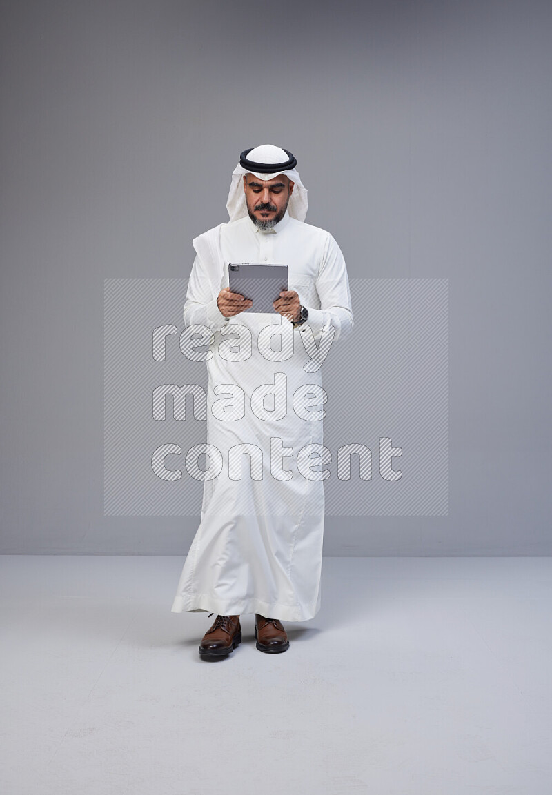 Saudi man Wearing Thob and white Shomag standing working on tablet on Gray background