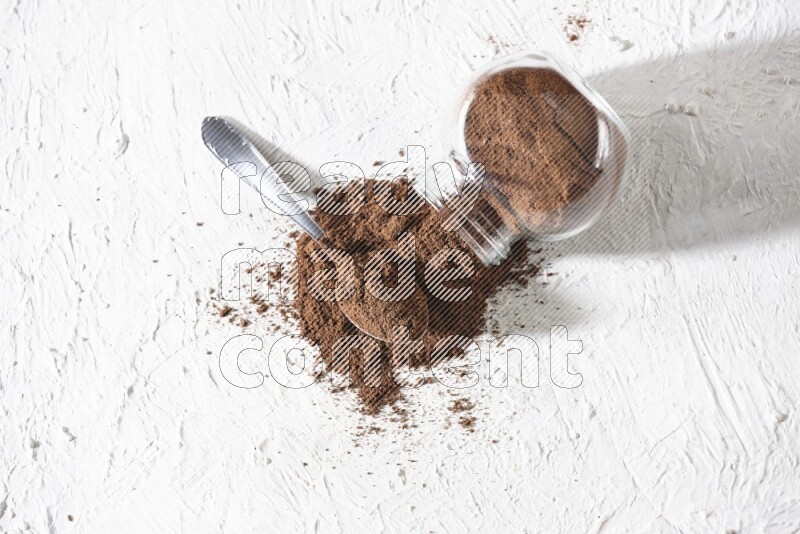 A flipped glass spice jar and a metal spoon full of cloves powder and powder came out of the jar on textured white flooring