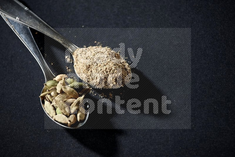 2 Metal spoons full of cardamom powder and cardamom seeds on black flooring