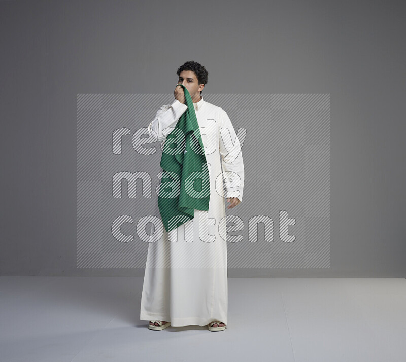 A Saudi man standing wearing thob kissing Saudi flag on gray background