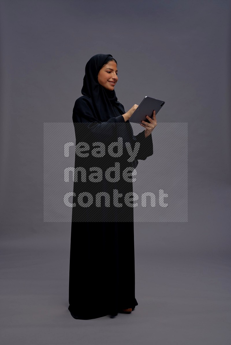 Saudi woman wearing Abaya standing working on tablet on gray background