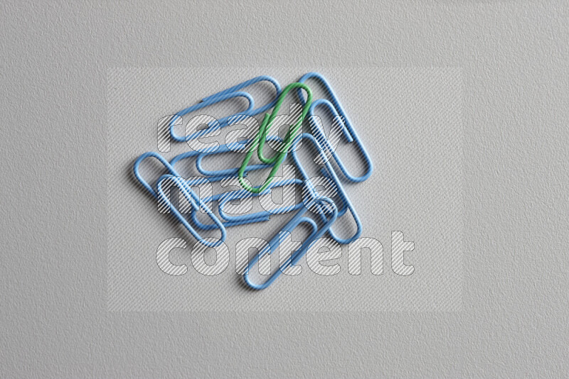 A green paperclip surrounded by bunch of blue paperclips on grey background
